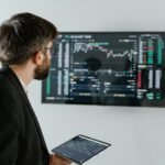 A businessman examines stock market data displayed on a monitor, holding a tablet.